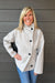 Woman wearing Tribal Sportswear Lofty Chic Funnel Neck Jacket In Antique White with jeans on a dark wood background.