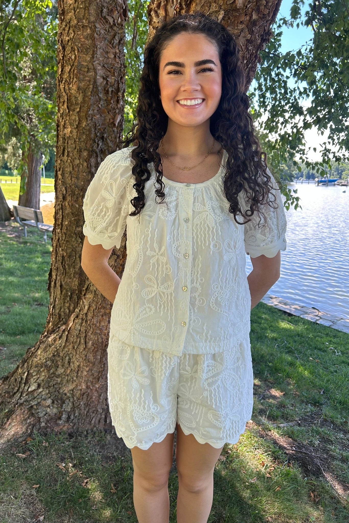 Woman in a light-colored outfit standing by a tree by a body of water