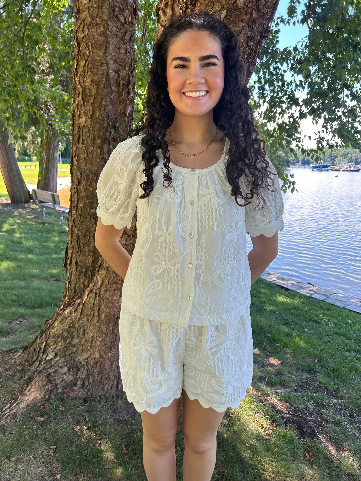 Woman in a light-colored outfit standing by a tree by a body of water