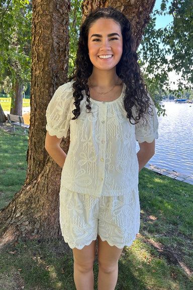 Woman in a light-colored outfit standing by a tree by a body of water