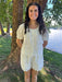 Woman in a light-colored outfit standing by a tree by a body of water