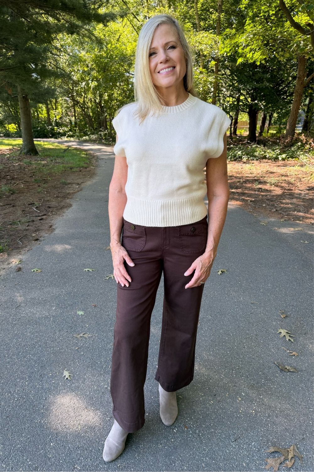 A woman standing on an outdoor paved path wearing a cream ribbed short-sleeve knit top, dark brown wide-leg pants, and light gray boots.