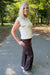 A woman with blonde hair stands on a paved path in a wooded outdoor setting, wearing a cream sleeveless knit top and dark brown wide-leg pants.