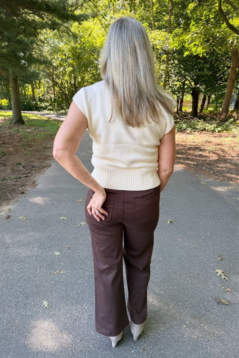 A woman viewed from behind wearing a cream short-sleeve ribbed knit top and dark brown wide-leg trousers, standing on an outdoor paved path surrounded by trees.