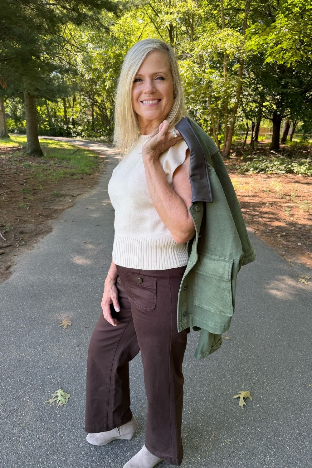A woman stands on a paved path in a wooded outdoor setting, wearing a cream ribbed sleeveless top, dark brown wide-leg pants, and holding an olive green jacket over her shoulder.
