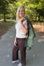 A woman stands on a paved path in a wooded outdoor setting, wearing a cream ribbed sleeveless top, dark brown wide-leg pants, and holding an olive green jacket over her shoulder.