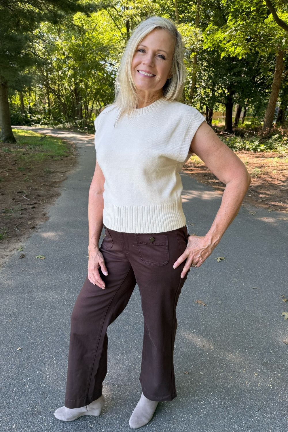 A smiling blonde woman stands on a paved path in a wooded outdoor setting, wearing a cream sleeveless ribbed knit top and dark brown wide-leg pants.
