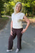 A smiling blonde woman stands on a paved path in a wooded outdoor setting, wearing a cream sleeveless ribbed knit top and dark brown wide-leg pants.