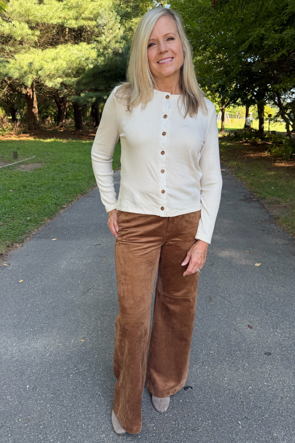 A woman standing on an outdoor paved path wearing a cream long-sleeve button-front cardigan and brown wide-leg corduroy pants.