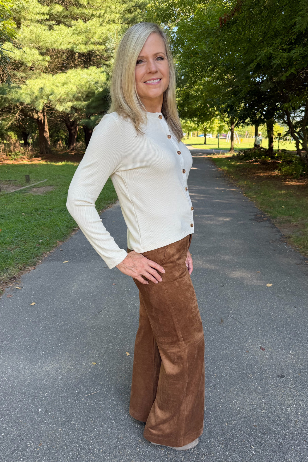 A woman stands on a paved path in a park, wearing a cream long-sleeve button-front top and brown wide-leg pants.