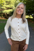 A smiling blonde woman wearing a cream-colored long-sleeve button-up top and brown pants, standing outdoors on a paved path with trees in the background.