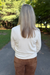 A woman viewed from behind wearing a cream-colored long-sleeve knit top and brown pants, standing on a paved path in a park with green trees in the background.