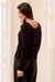 A woman wearing a black long-sleeve knit top with decorative gold buttons down the back, shown from a side-back angle against a soft cream curtain background.