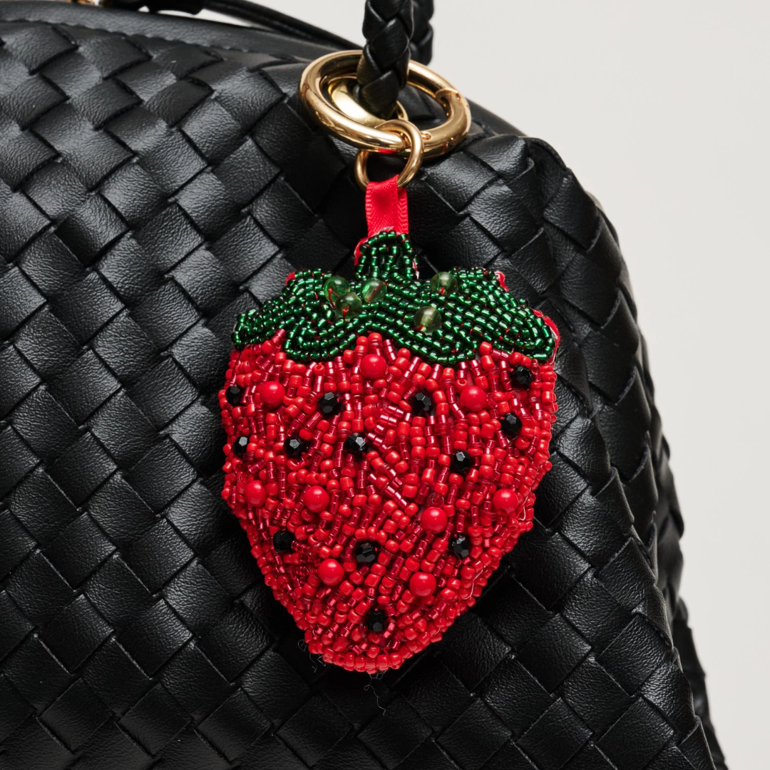A red and green beaded strawberry charm with black seed details hanging from a gold ring on a black woven leather bag, photographed close-up against a light gray background.