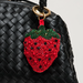 A red and green beaded strawberry charm with black seed details hanging from a gold ring on a black woven leather bag, photographed close-up against a light gray background.