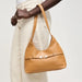 A tan leather hobo shoulder bag with a front strap detail and silver-tone hardware, carried by a model wearing cream pants against a light gray studio background.
