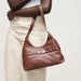 A brown leather hobo shoulder bag with a front strap detail and gold-tone hardware, shown being worn by a model in cream pants against a light gray studio background.