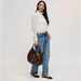 A woman stands against a light gray studio background wearing a white lace long-sleeve blouse, blue jeans, black flats, and holding a brown leather hobo bag.