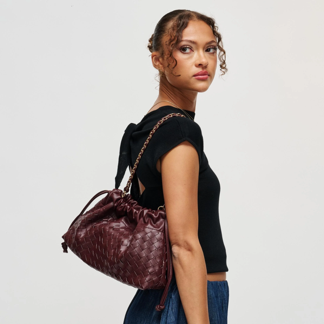 A woman in a black top and blue jeans carries a dark burgundy woven leather shoulder bag with a gold chain strap against a light gray studio background.