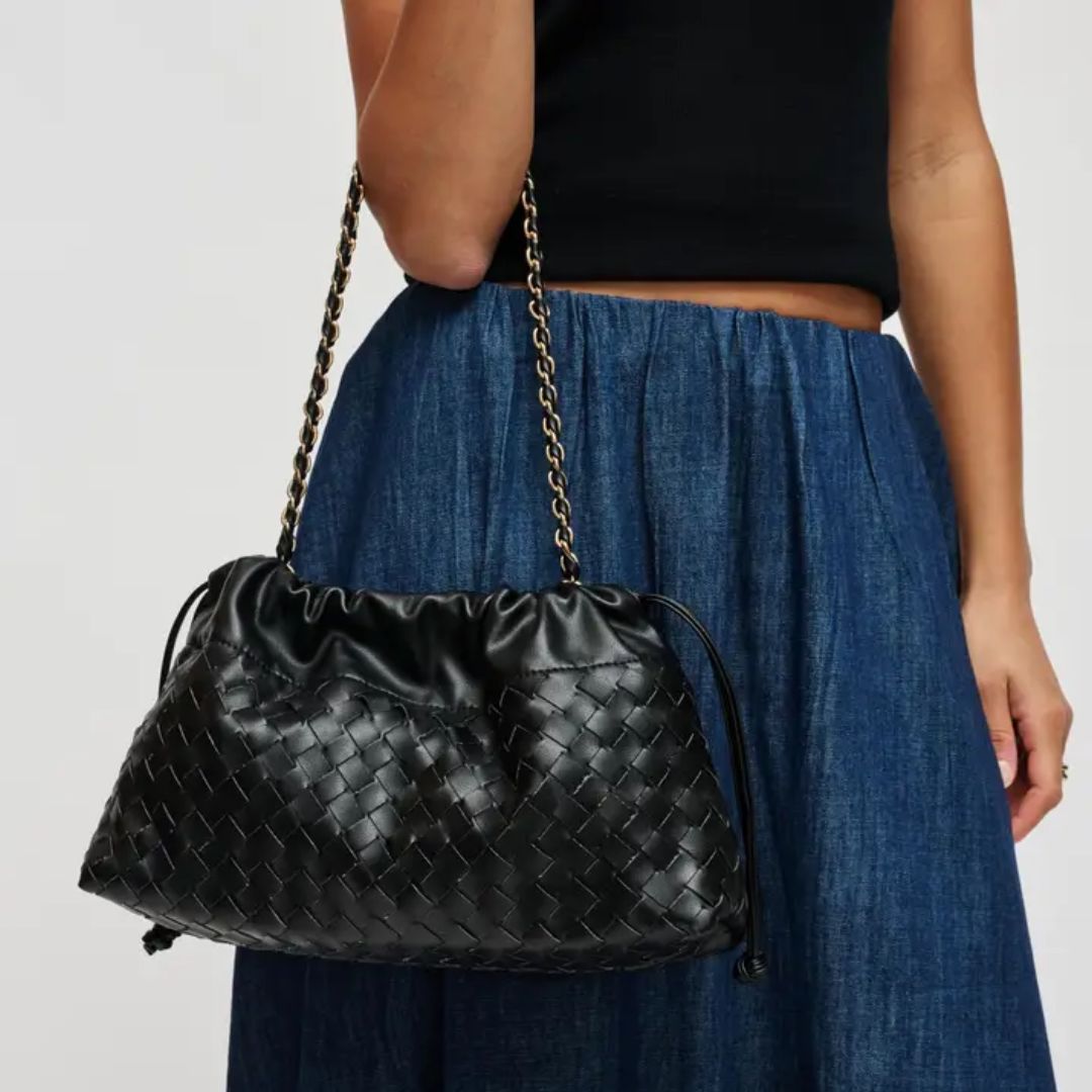 A black woven leather shoulder bag with a gold chain strap, worn by a model wearing a black top and a blue pleated skirt against a light gray background.