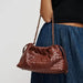 A brown woven leather shoulder bag with a chain strap, worn by a model wearing a black top and blue denim skirt against a light gray background.