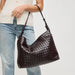 A dark brown woven leather shoulder bag with a slouchy hobo shape, carried on the shoulder of a person wearing a white top and light blue jeans against a light gray background.