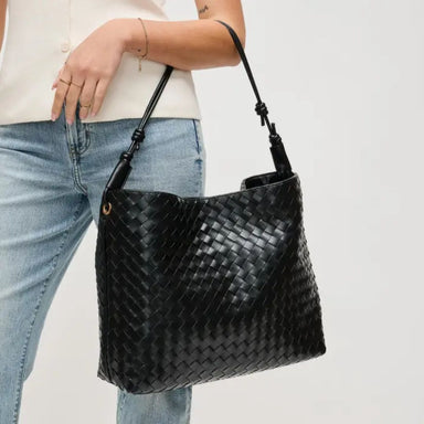 A black woven leather shoulder bag with a top handle, carried by a model wearing a cream top and light blue jeans against a plain white background.