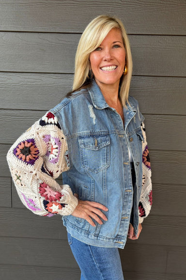 A smiling blonde woman wearing a light blue denim jacket with colorful crocheted granny-square sleeves, posed against a dark gray wood-paneled wall.