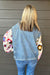 A person viewed from behind wearing a light blue denim jacket with colorful crocheted sleeves, paired with blue jeans, standing against a dark gray wood-paneled wall.