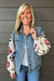 A smiling blonde woman wearing a light blue denim jacket with colorful crocheted granny-square sleeves, posed against a dark gray wood-paneled wall.