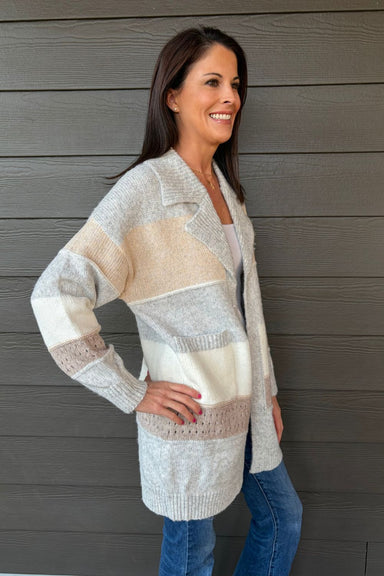 Woman wearing Very Moda Chunky Colorblock Cardigan In Gray with jeans on a dark wood background.