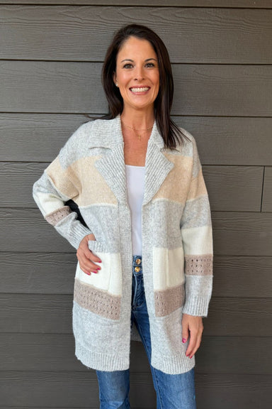 Woman wearing Very Moda Chunky Colorblock Cardigan In Gray with jeans on a dark wood background.