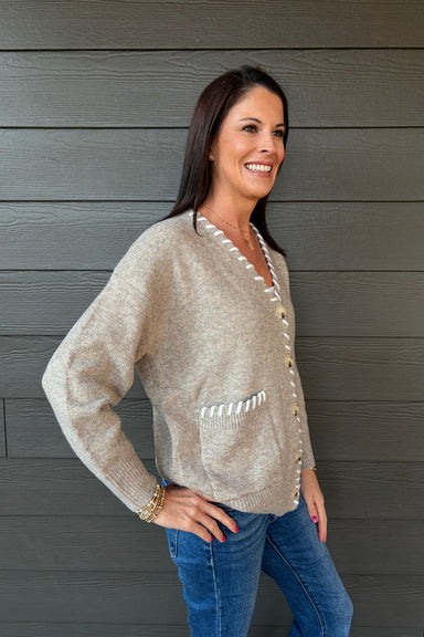 Woman wearing Very Moda Contrast Whipstitch Cardigan In Khaki with jeans on a dark wood background.