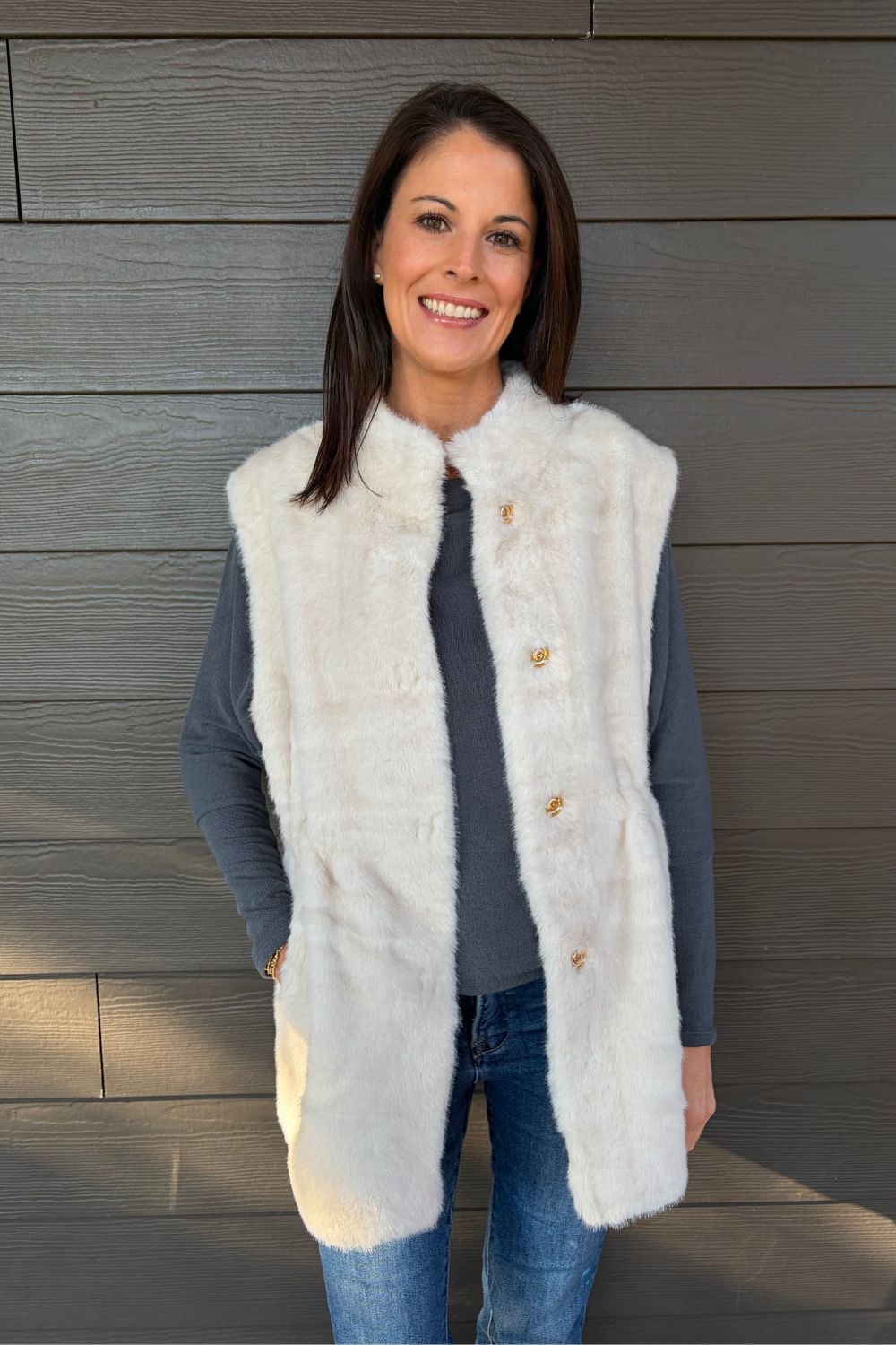 Woman wearing Very Moda Fur Snap Vest In White with jeans on a dark wood background.