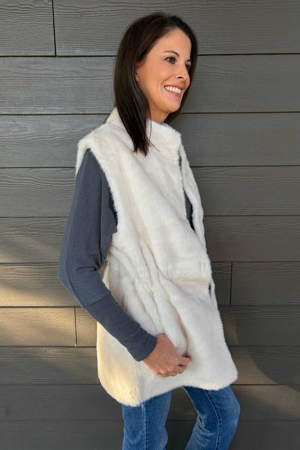 Woman wearing Very Moda Fur Snap Vest In White with jeans on a dark wood background.