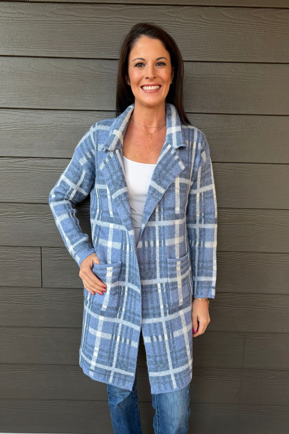 A woman wearing a blue and white plaid coat over a white top and blue jeans, standing against a dark gray wood-paneled wall.