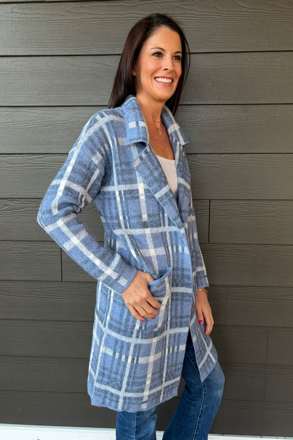 A woman wearing a blue and white plaid coat over a white top and blue jeans, standing against a dark gray wood-paneled wall.