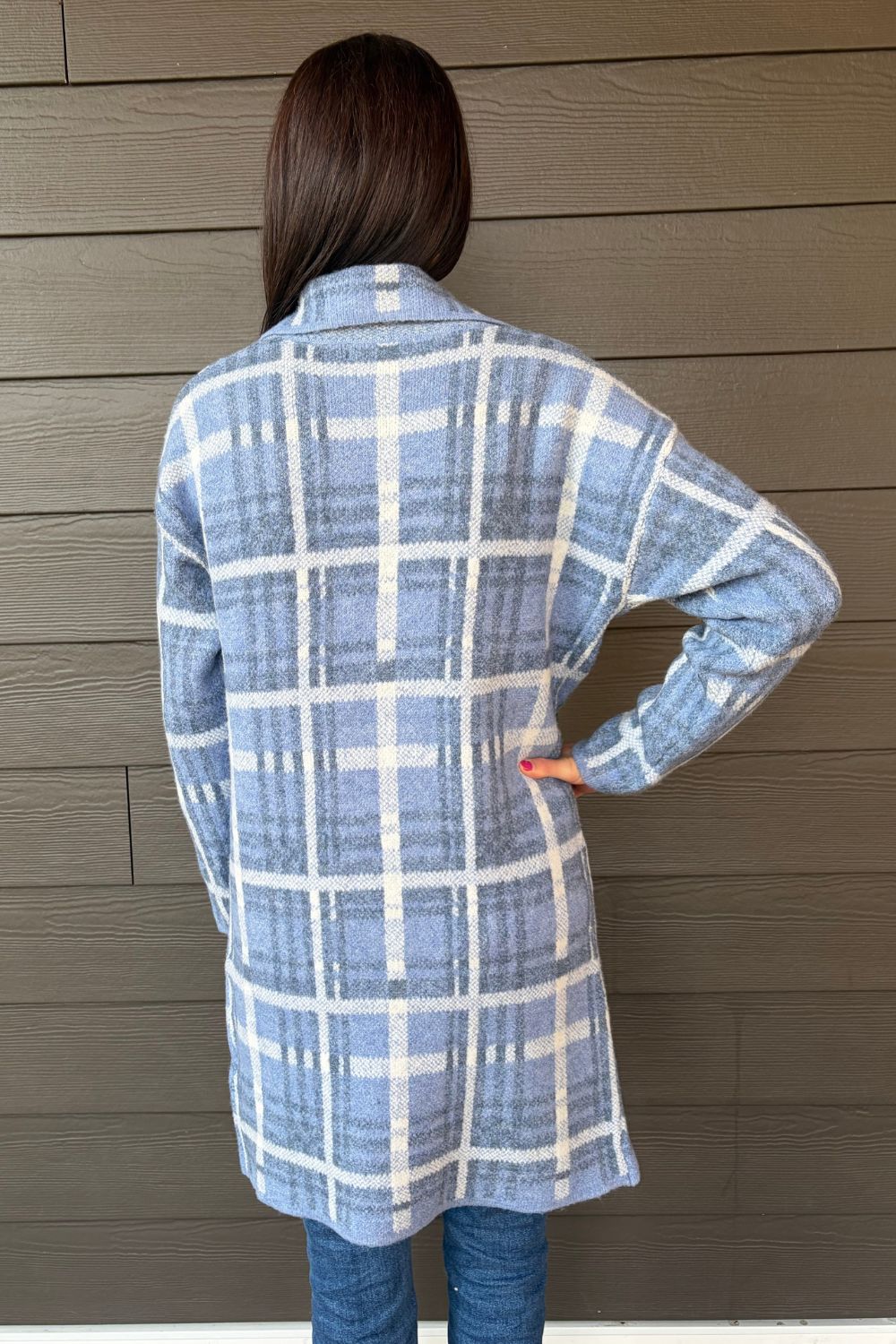 A person shown from the back wearing a light blue and white plaid flannel shirt jacket, standing against a brown horizontal siding wall.