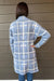 A person shown from the back wearing a light blue and white plaid flannel shirt jacket, standing against a brown horizontal siding wall.