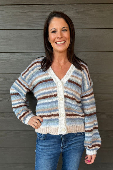 A woman wearing a striped knit cardigan sweater with blue jeans, standing against a dark gray wood-paneled wall.