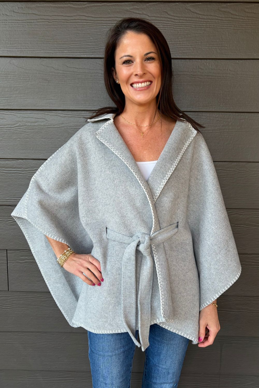 Woman wearing Very Moda Whipstitch Belted Cape In Gray with jeans on a dark wood background.