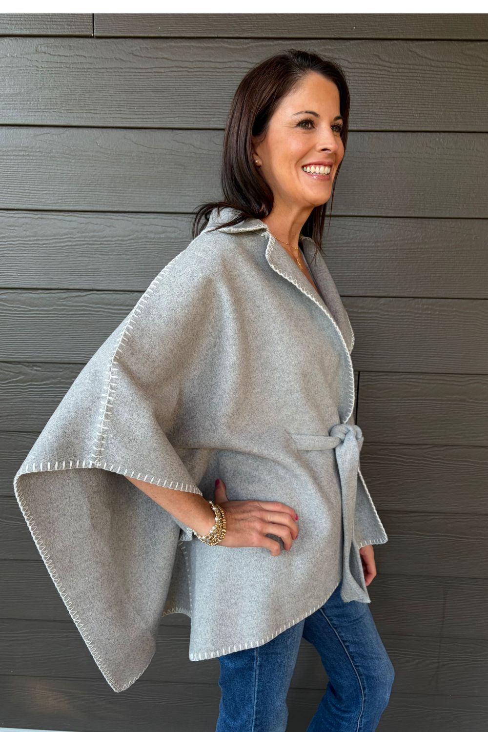 Woman wearing Very Moda Whipstitch Belted Cape In Gray with jeans on a dark wood background.