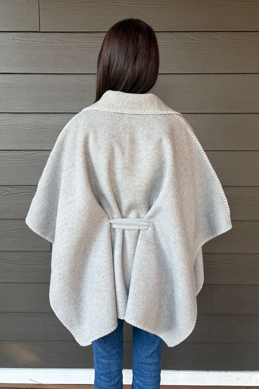 Woman wearing Very Moda Whipstitch Belted Cape In Gray with jeans on a dark wood background.
