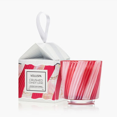 A Voluspa Coconut Wax Candle in a red and pink striped glass jar, shown next to its white and red patterned gift box with a white hanging ribbon on a clean white background.