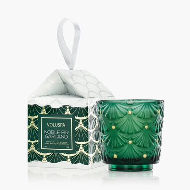A green faceted glass candle holder with gold leaf-like accents stands next to its patterned gift box with a white hanging ribbon. The box is labeled VOLUSPA NOBLE FIR GARLAND.