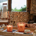 Two lit candles in decorative holders on a wooden tray with a stone wall and outdoor view in the background.