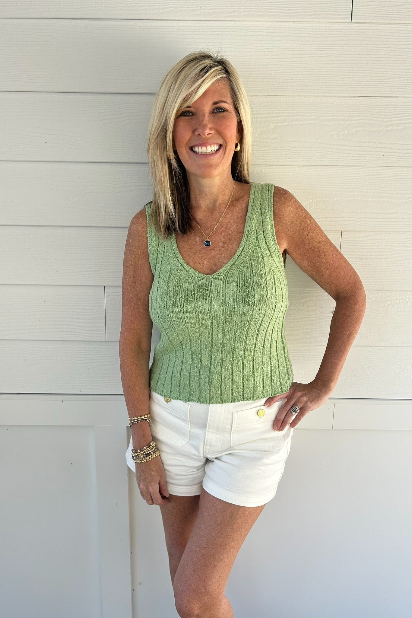 Woman wearing a green sleeveless top and white shorts standing against a white wooden background