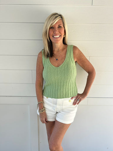 Woman wearing a green sleeveless top and white shorts standing against a white wooden background