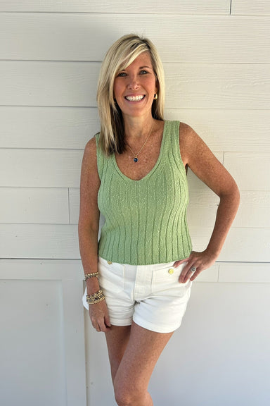 Woman wearing a green sleeveless top and white shorts standing against a white wooden background
