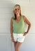 Woman wearing a green sleeveless top and white shorts standing against a white wooden background